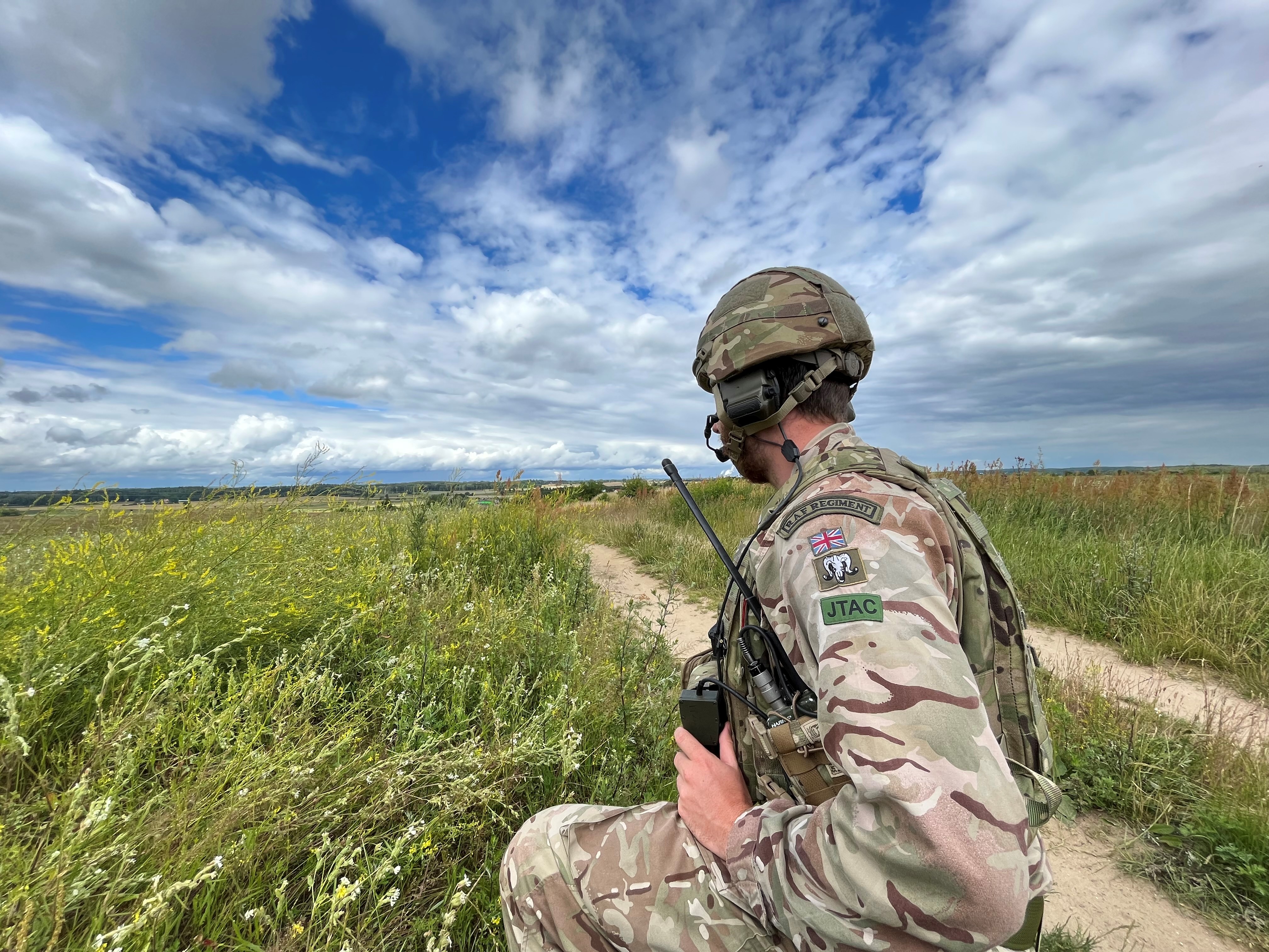 Image shows RAF Regiment crouching with radio, in a grassy landscape.